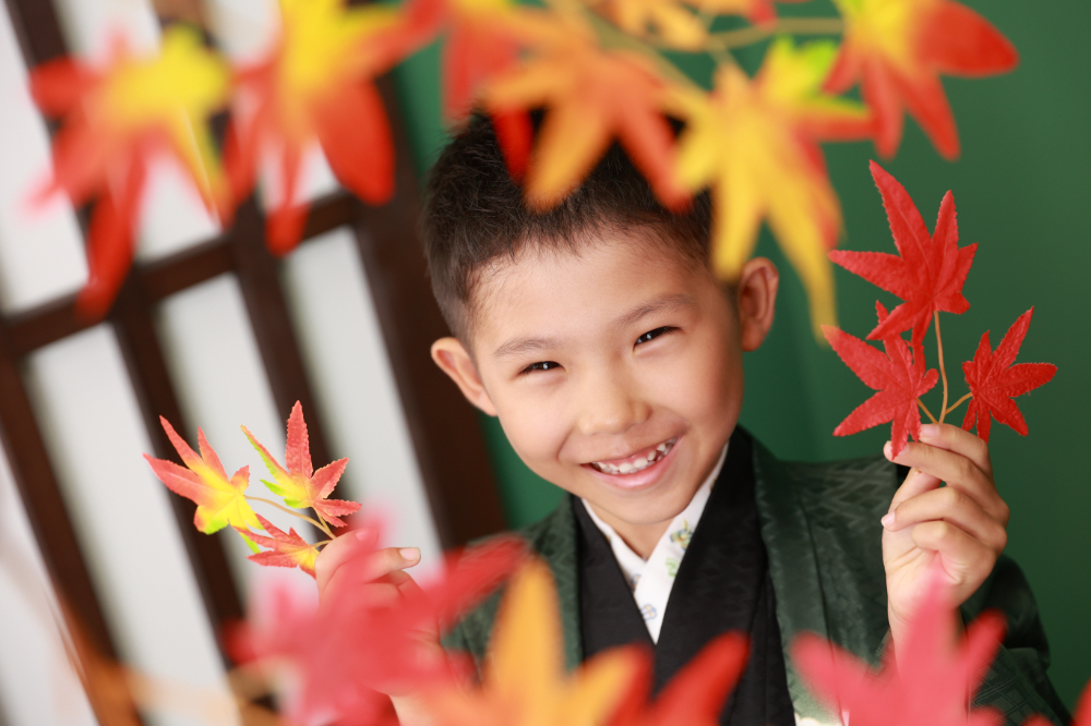 紅葉越しに笑顔を見せる緑の着物の5歳男の子