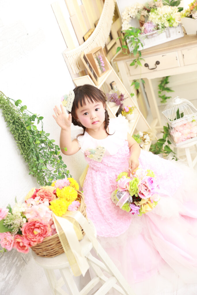 ピンクのドレスと花に囲まれた3歳の女の子がブーケを持つ姿