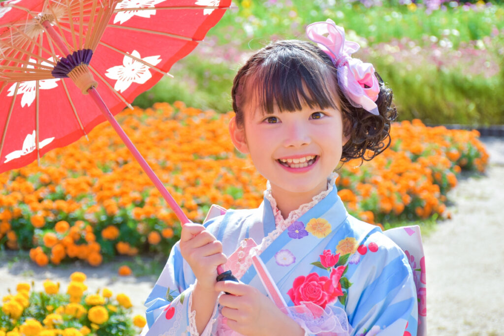 水色の着物を着た7歳の女の子が、花壇の前でポーズ